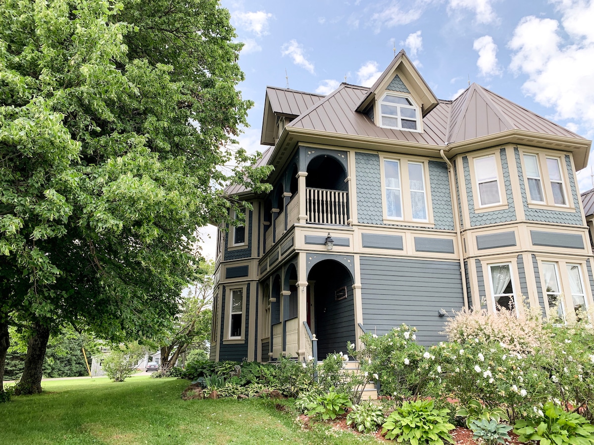 A historic heritage home is displayed, showcasing intricate architectural details and a mixture of blue and cream siding. The entrance features a small balcony, framed by large windows. Surrounding greenery adds to the appealing setting.