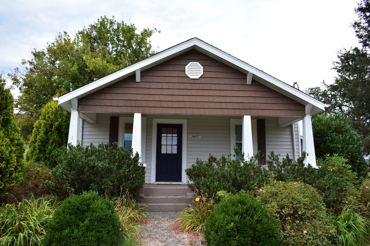 The exterior of the farmhouse is showcased, featuring a symmetrical structure with a brown shingle roof. A dark blue door is centered between two white columns, framed by lush greenery and manicured bushes that create a welcoming approach.