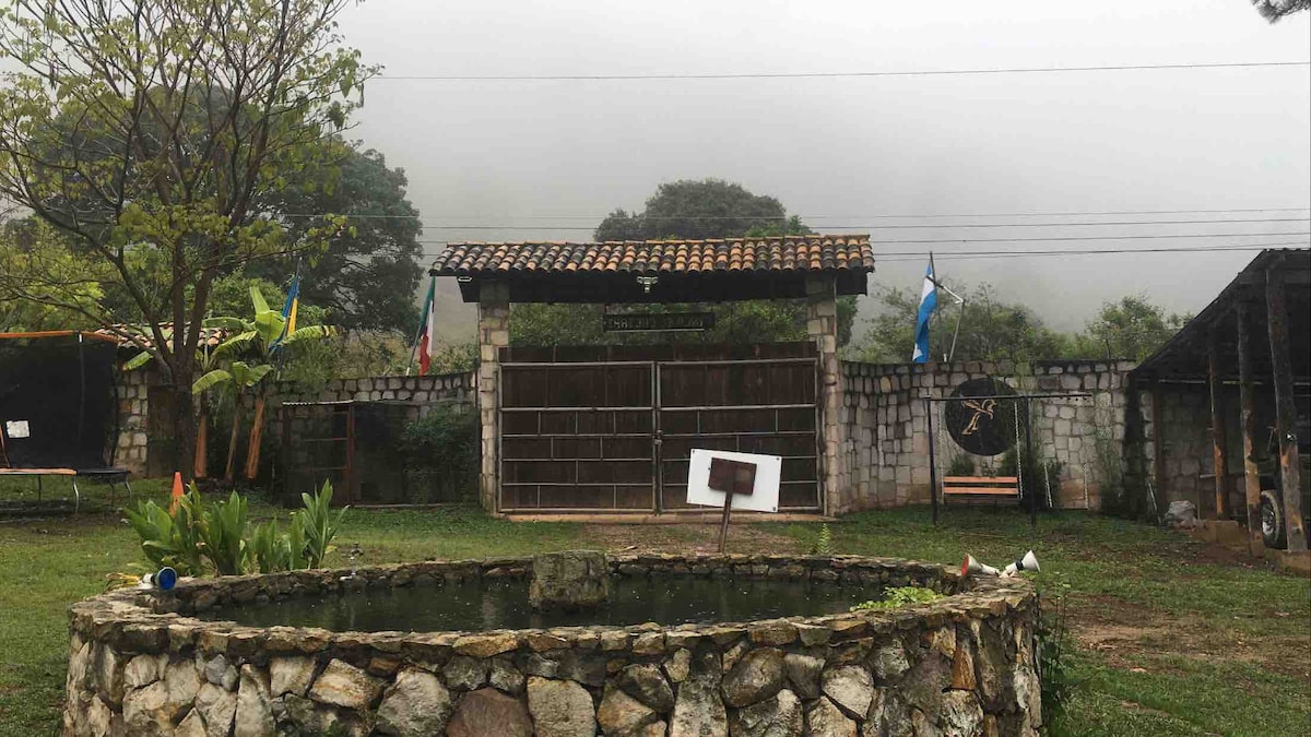A serene entrance to the property is framed by a rustic stone wall and wooden gate, surrounded by lush greenery. Fog blankets the background, enhancing the peaceful atmosphere. A decorative pond sits in the foreground, adding to the natural charm of the setting.