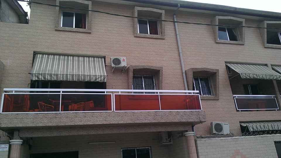 A multi-story building features a light-colored brick exterior with multiple windows. The lower level showcases a balcony with orange-tinted glass railings, and the upper floors have striped awnings above the windows. Air conditioning units are present on the facade.