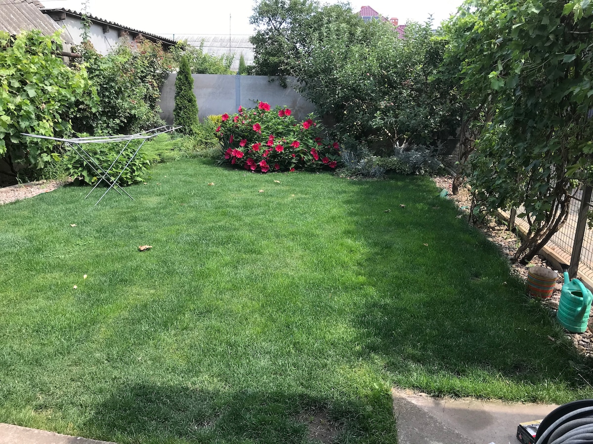 A well-maintained garden area is featured, showcasing a lush green lawn. Vibrant pink flowers are located along one side, surrounded by various shrubs and greenery. The space appears inviting, with a clothesline positioned nearby. A fence and trees create a sense of privacy.