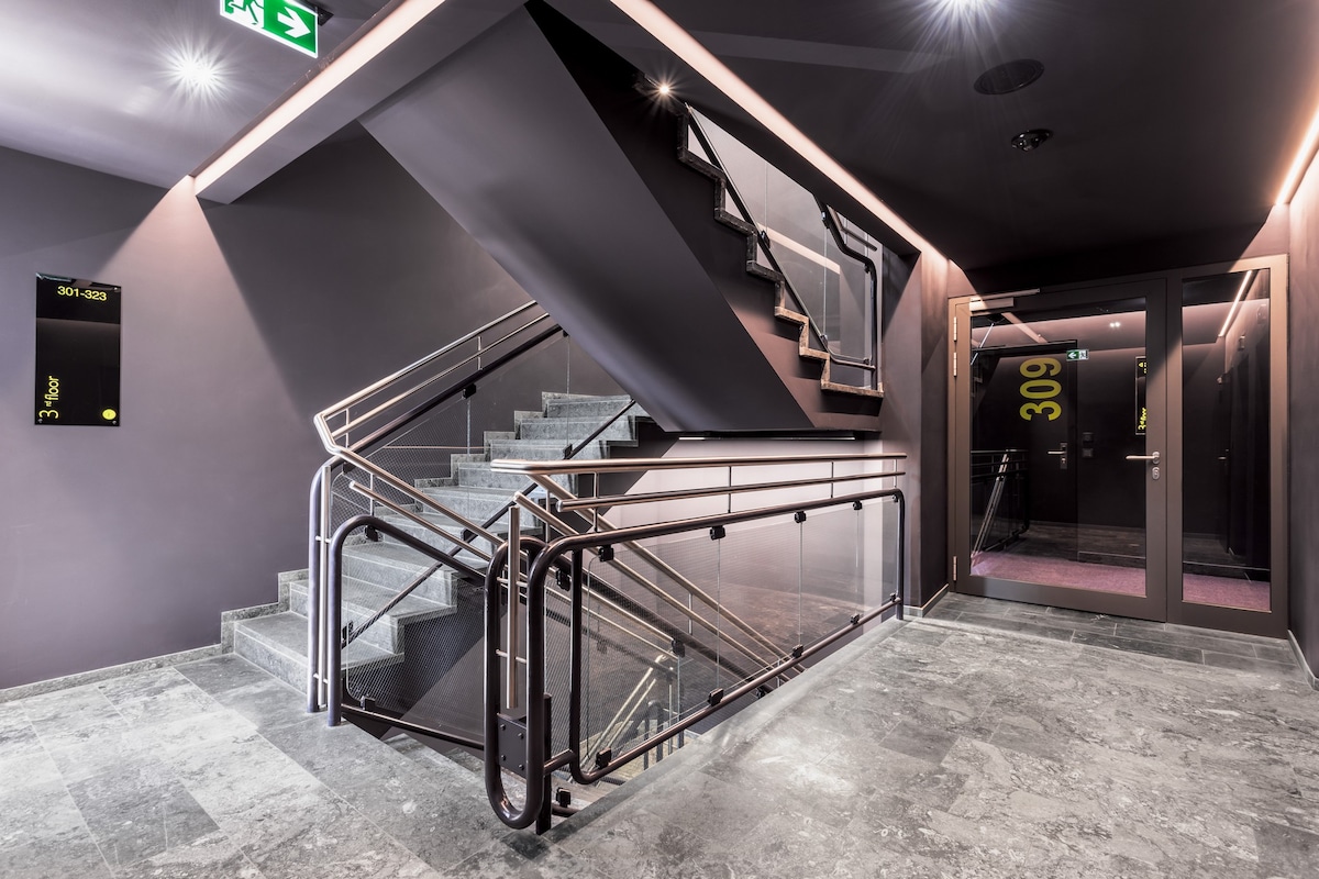 A modern staircase with a sleek metal railing is illuminated by soft lighting. Dark walls create a sophisticated ambiance. The polished marble floor reflects the lighting, while clear signage is visible on the walls for ease of navigation to the upper floors.