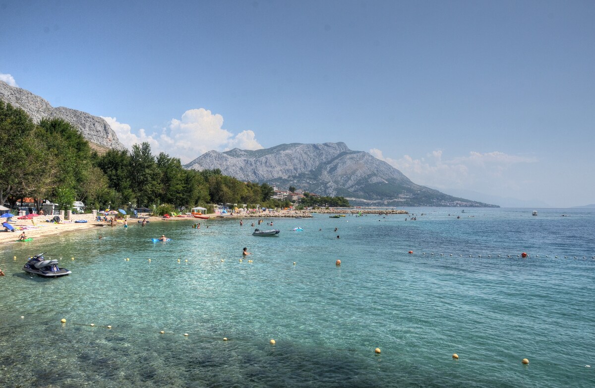 A calm beach scene is displayed, featuring clear, shallow waters with a sandy shoreline. Visitors are seen enjoying recreational activities, while the backdrop includes gentle hills and mountains under a clear blue sky.
