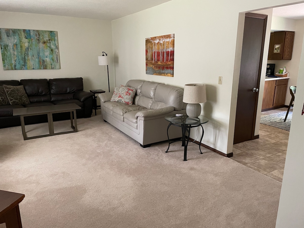 A spacious living room is depicted, featuring a light-colored carpet and neutral walls. A comfortable sectional sofa in a dark hue and a light gray sofa are arranged around a coffee table. A lamp and a small side table accompany the seating, with a glimpse of the kitchen in the background.