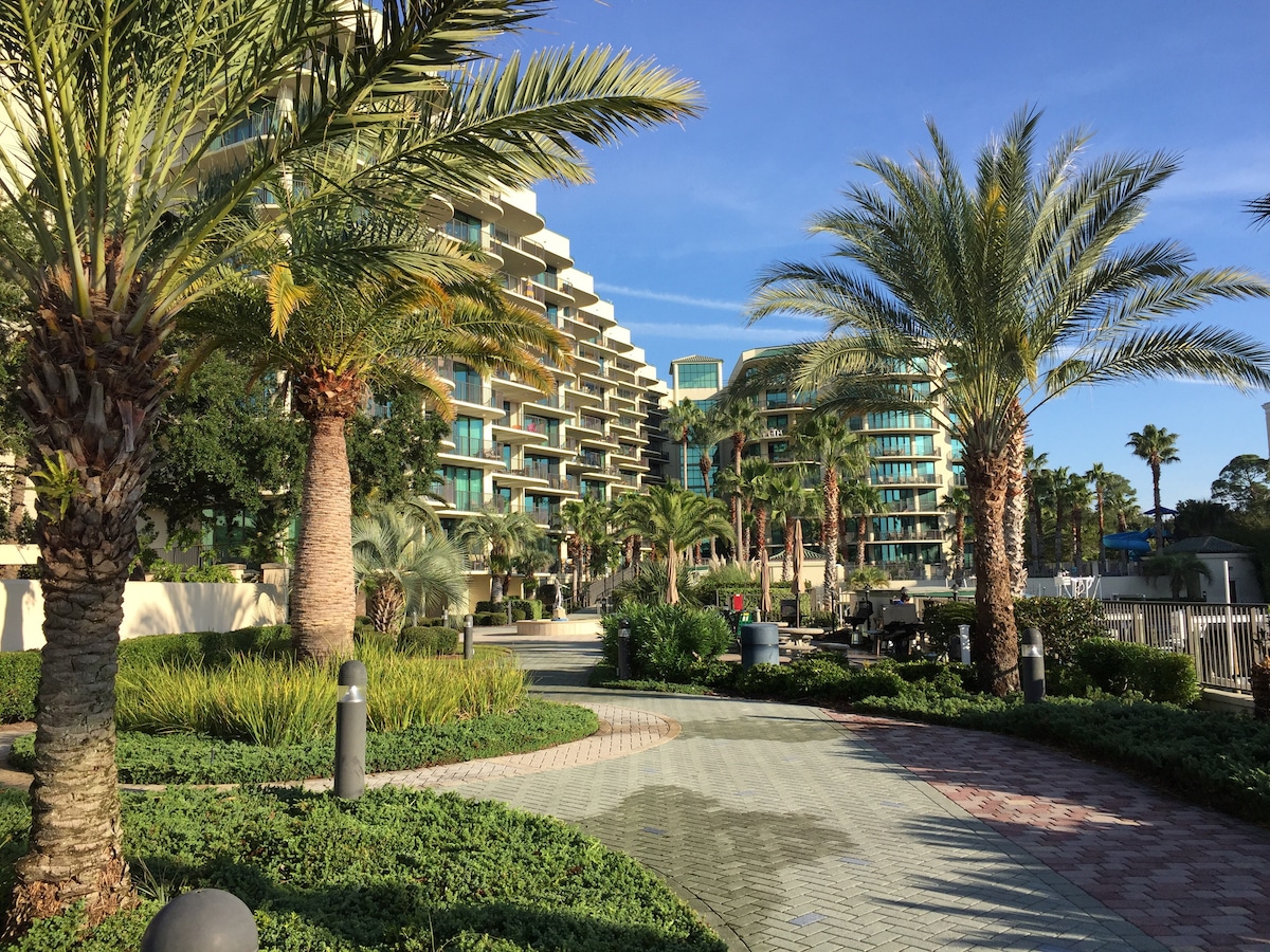 A landscaped pathway is bordered by lush palm trees and greenery, leading towards a modern multi-story building with balconies. Sunlight casts gentle shadows on the paved walkway, creating a serene atmosphere. Outdoor seating and gentle curves of the path enhance the inviting environment.