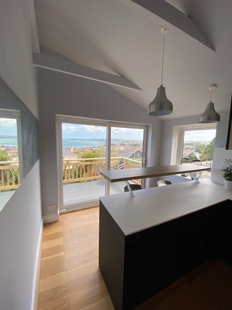 The kitchen features a modern design with a white countertop and dark cabinets. Large windows allow ample natural light and showcase views of the sea and surrounding area. A wooden dining table is positioned near the windows, inviting gatherings with scenic backdrops.