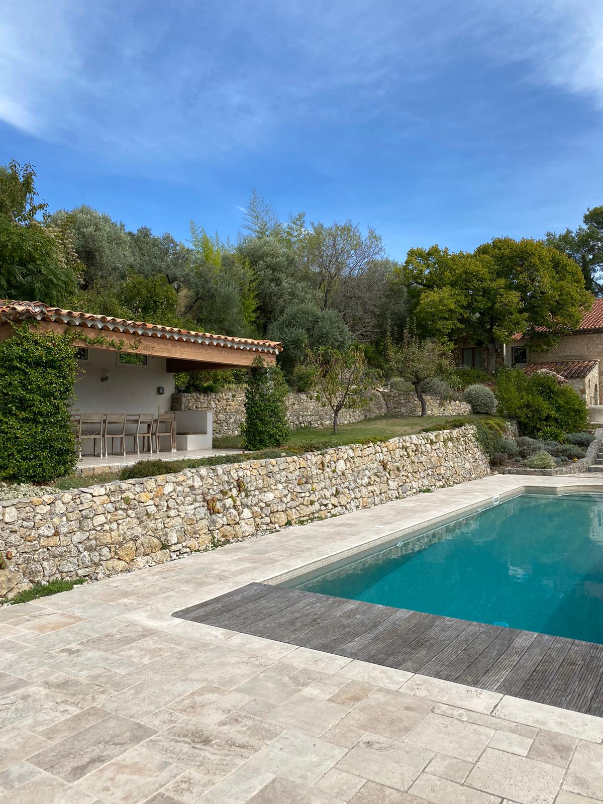 A pool area features a clear blue swimming pool set within a stone-surrounded terrace. Adjacent to the pool, a shaded pool house offers seating and a bar arrangement, surrounded by lush greenery and terraced landscaping, creating a serene outdoor environment.