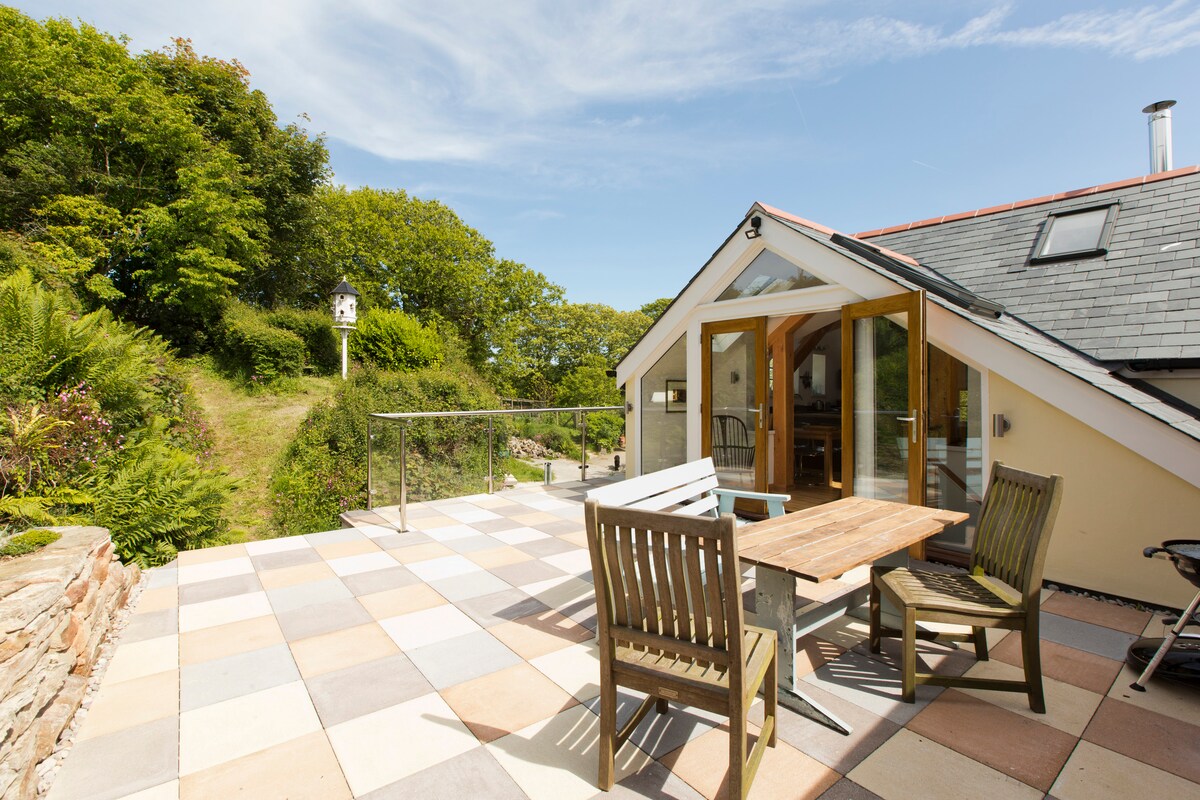 An outdoor patio area features a wooden dining table surrounded by garden chairs. The glass balustrade offers unobstructed views of the surrounding greenery. Natural light illuminates the space, enhancing the inviting atmosphere for outdoor meals or relaxation.
