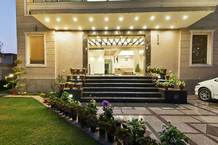 An elegant entrance is framed by well-maintained greenery and flower pots lining the pathway. Steps lead up to a spacious lobby area, illuminated by decorative lights. The modern facade features large windows that invite natural light into the interior spaces.