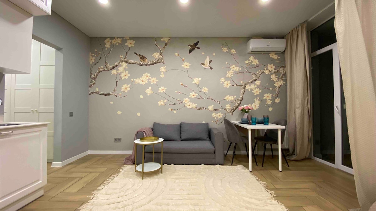 A compact sofa sits against a floral wallpaper accented by birds, contributing to a serene ambiance. A small dining table is nearby, flanked by elegant chairs. Soft lighting and a rug enhance the comfort of the space, which features light-colored walls and a window allowing natural light to enter.