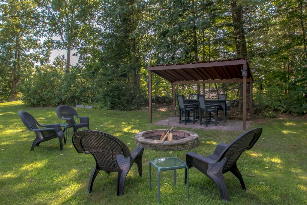 An outdoor seating area is arranged around a stone fire pit, with several black chairs positioned for comfort. A shaded pavilion contains a dining table, surrounded by greenery, providing a peaceful space for gatherings or relaxation.