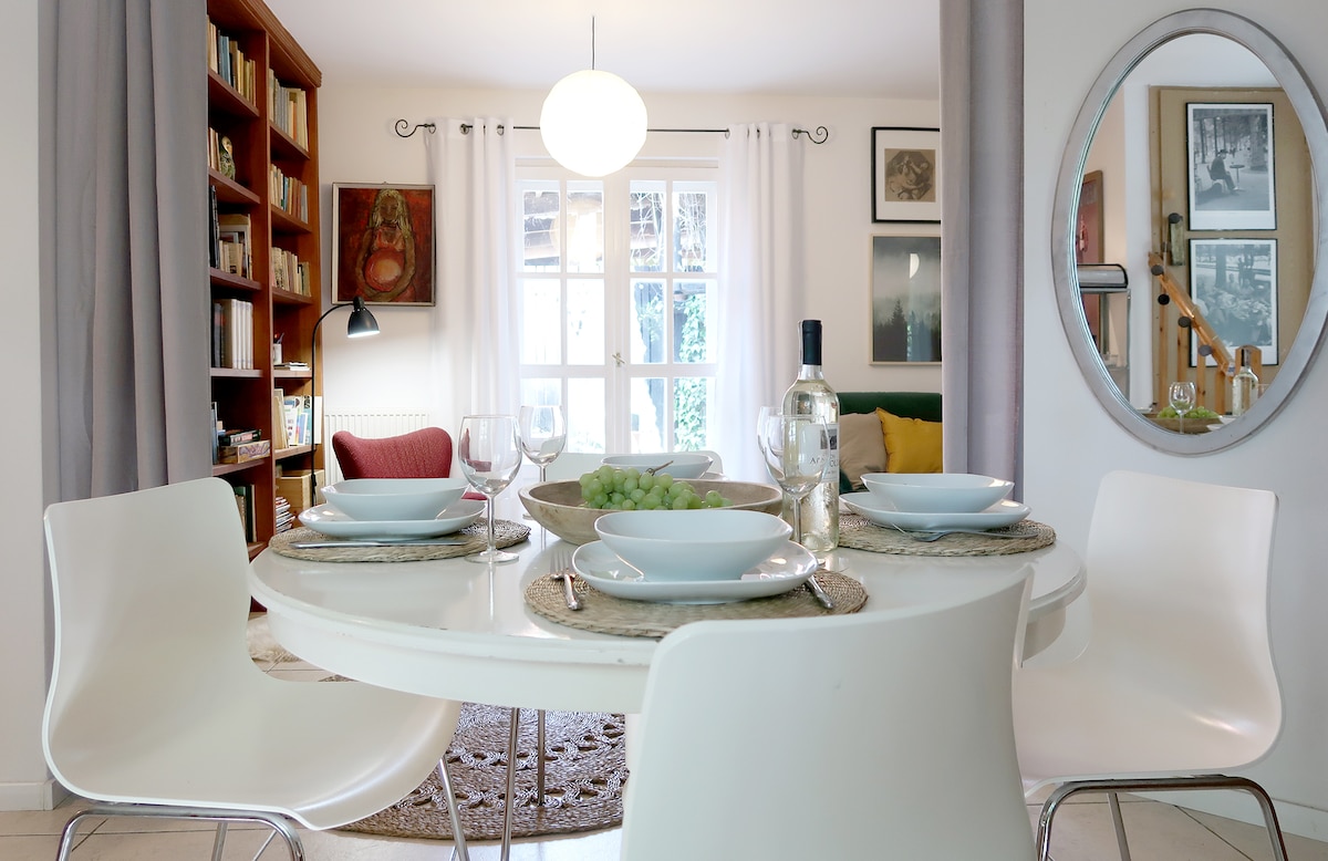 A dining area features a round table set with plates and wine glasses, accompanied by a bowl of grapes. A stylish white bookshelf lines one wall, while a comfortable living space is visible in the background, highlighted by a large window and various artworks.