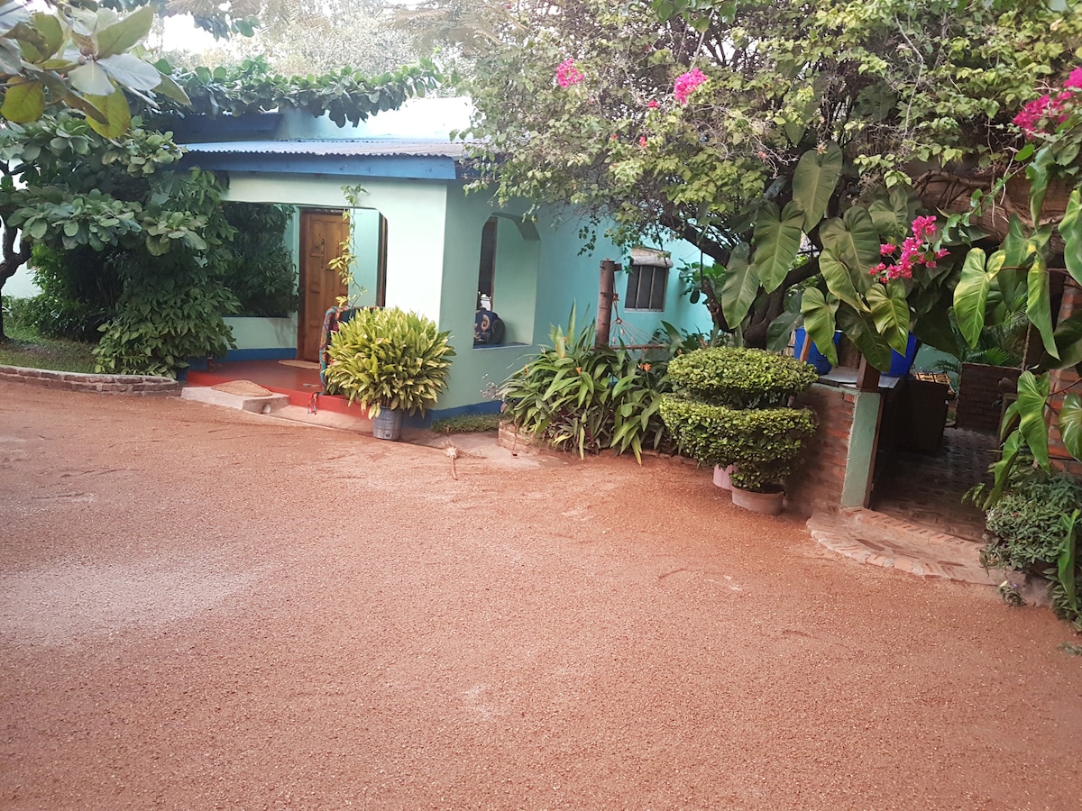 A charming cottage is nestled among lush greenery, with vibrant flowering plants framing the entrance. The light blue exterior is complemented by a red pathway leading to a welcoming door. A well-maintained garden area is visible, enhancing the tranquil outdoor setting.
