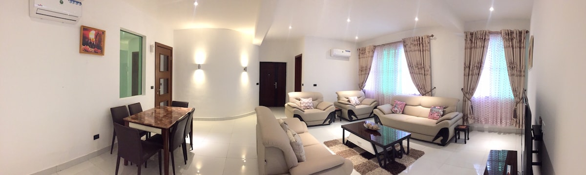 A spacious living area is presented with comfortable seating arrangements, including a large sofa set and armchairs. Natural light filters through two windows adorned with sheer curtains. A dining table is visible, accompanied by four chairs, situated next to an accent wall featuring artwork.
