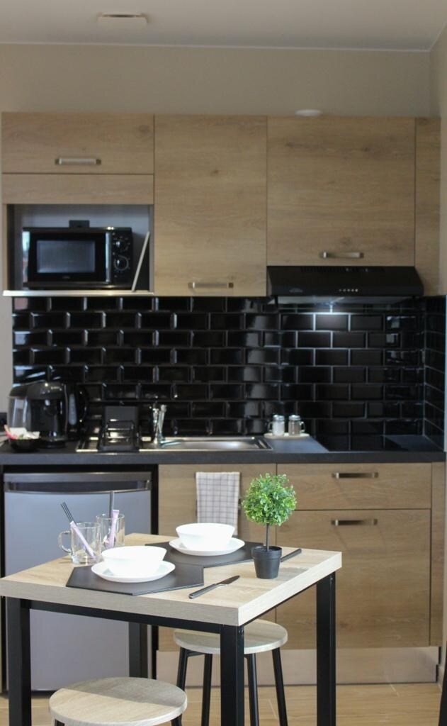 A compact kitchen area features modern cabinetry in warm wood tones, complemented by sleek black tile backsplash. Essential appliances include a microwave and coffee machine. A small dining table is set with white dishware and a decorative plant, providing a cozy space for meals.