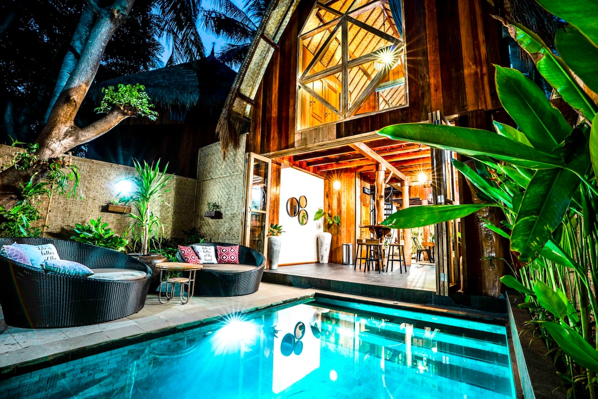 An outdoor area is showcased with seating arrangements featuring dark wicker chairs and a private plunge pool reflecting the surrounding lights. Tropical plants offer a lush backdrop, and the building's large windows reveal a warm interior space, enhancing the connection with nature.