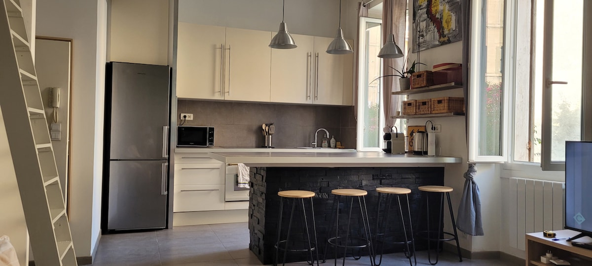A modern kitchen is illuminated by natural light from large windows, featuring white cabinetry and a sleek gray countertop. Four stools are positioned at a dark island, complementing stainless steel appliances and minimalist decor, creating a functional and inviting space for meal preparation.