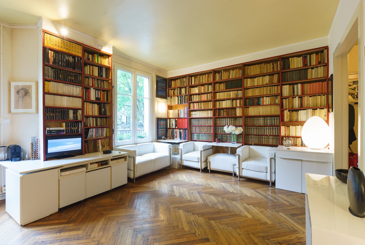 A well-organized living area features a large wall of shelves filled with books, complemented by two white armchairs and a modern coffee table. Natural light filters through large windows, illuminating a warm parquet floor, creating a bright and spacious environment.