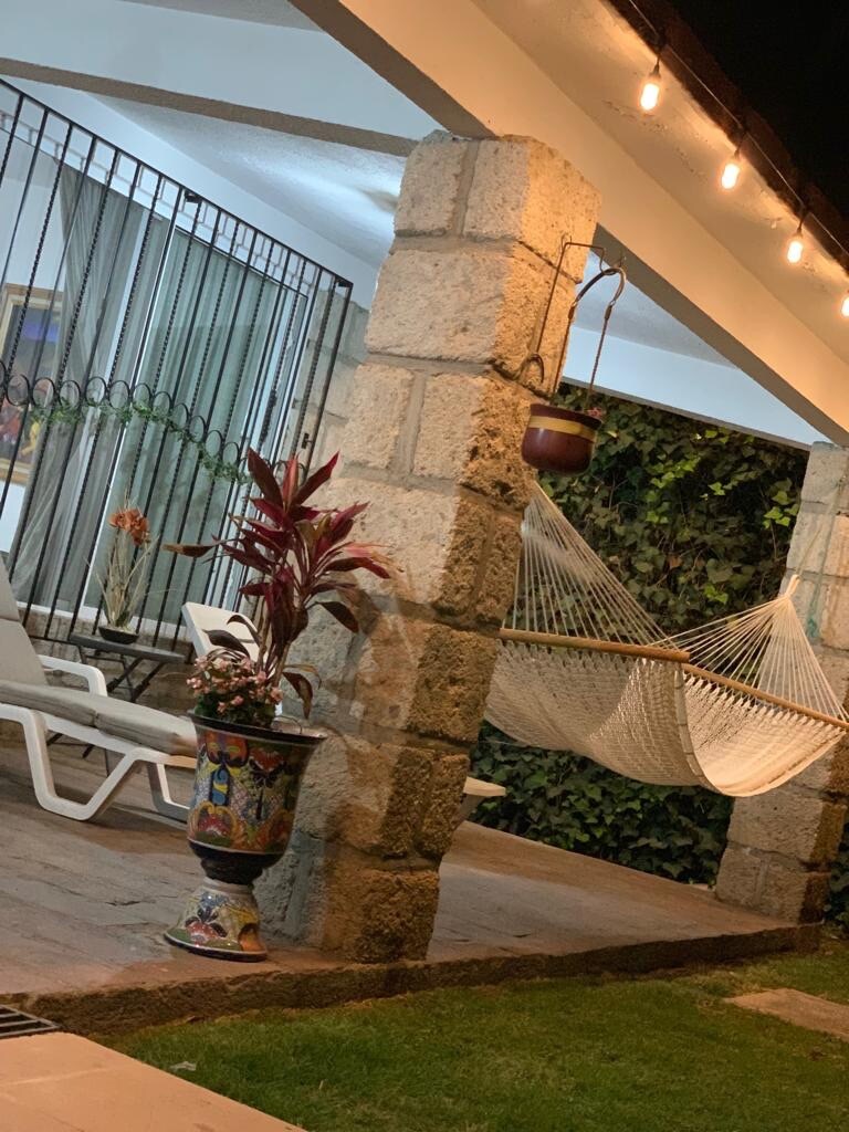 A serene outdoor area showcases stone columns supporting a shaded porch. A colorful planter holds a flowering plant, while a hammock is suspended between the columns. Sun loungers are arranged nearby, and string lights add a warm glow to the setting.