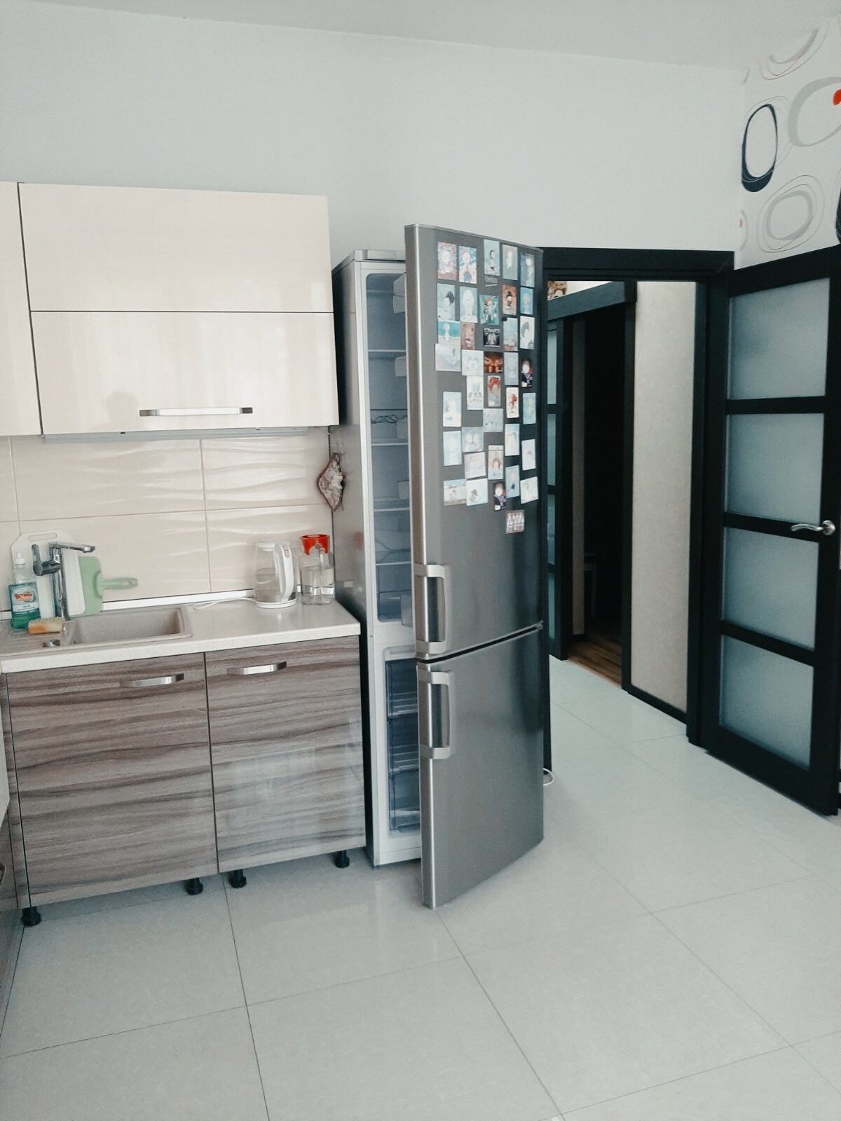 A modern kitchen is seen featuring a silver refrigerator with the door ajar, revealing organized shelves. The cabinetry combines light wood and sleek white finishes. A sink is situated adjacent to the refrigerator, while a partially open door leads to a hallway, contributing to the spacious feel.