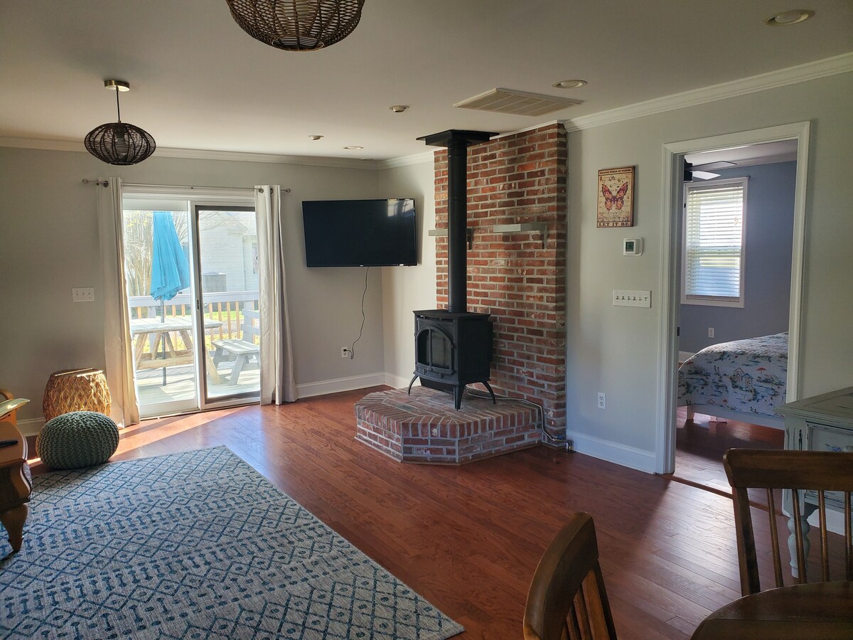 An open living space is highlighted by a brick fireplace and a large flat-screen television mounted on the wall. Natural light floods in through sliding glass doors, leading to an outdoor deck. A cozy rug and dining area enhance the comfortable atmosphere.