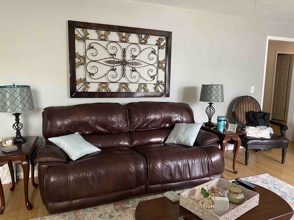 A spacious living room area features a brown leather couch adorned with two light blue cushions. Decorative wall art is displayed above the couch. Nearby, two lamps with patterned shades provide soft lighting, while a wooden side chair complements the arrangement.