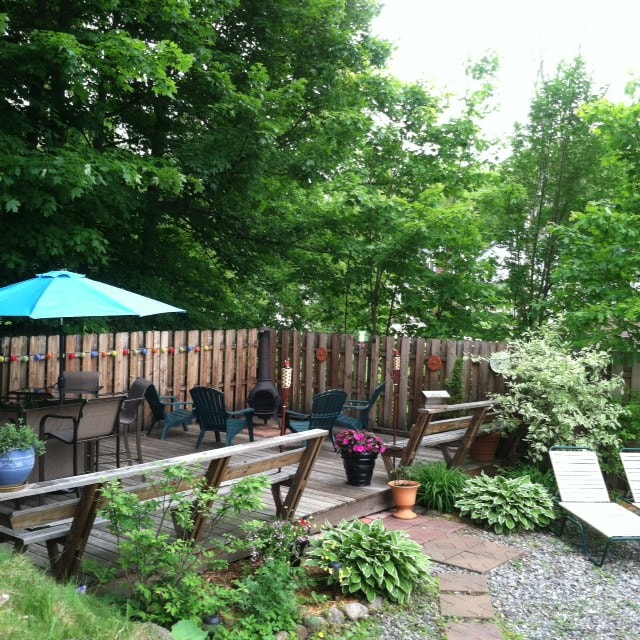 An outdoor patio area features a mix of seating options, including chairs and lounge chairs arranged around a gravel path. A wooden fence surrounds the space, complemented by vibrant greenery and flowering plants. An umbrella provides shade over a table, enhancing the outdoor experience.