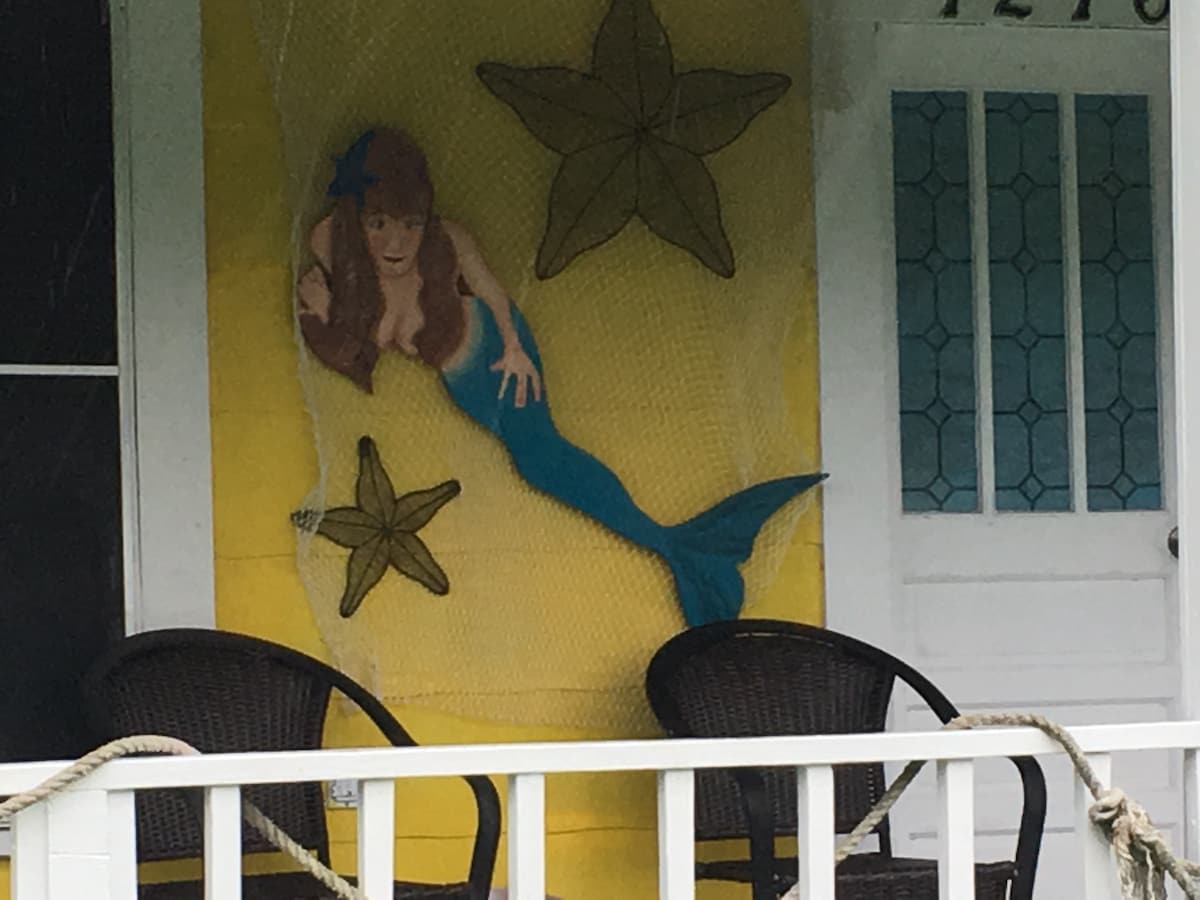 A vibrant yellow wall features a decorative mermaid and starfish artwork, set against a fishing net. Two wicker chairs are positioned on a covered porch, providing a cozy seating area for relaxation.