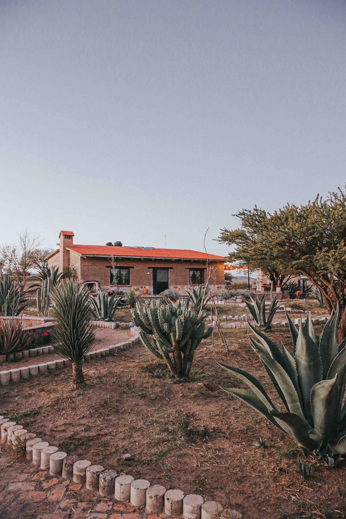 A charming residence features a warm-toned exterior with a red-tiled roof. Surrounding gardens showcase a variety of cacti and lush greenery, creating an inviting outdoor space. A pathway lined with stones leads towards the house, enhancing the tranquil environment.