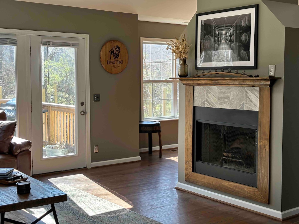 A cozy living area features a rustic fireplace framed by light-colored stone. Large windows allow natural light to fill the space, with a view of greenery outside. A framed photograph adorns the wall above the fireplace, and a wooden side table is positioned nearby.
