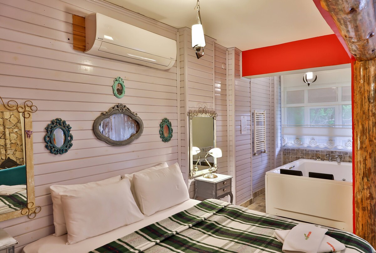 A cozy bedroom features a spacious bed with white linens and a plaid blanket. Mirrors are displayed on the walls, complemented by a modern air conditioning unit. Natural light enters from adjacent glass doors leading to a relaxing area with a bathtub.