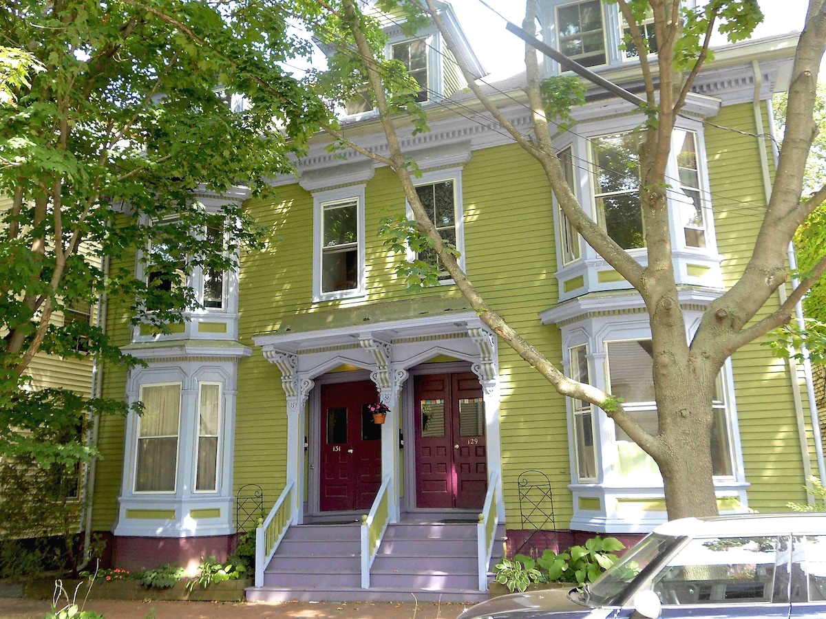 The front view of a classic Victorian duplex displays a vibrant green exterior with intricate architectural details. Two inviting entryways feature maroon doors, framed by large windows and decorative trim. Lush greenery surrounds the property, enhancing the welcoming atmosphere of the neighborhood.