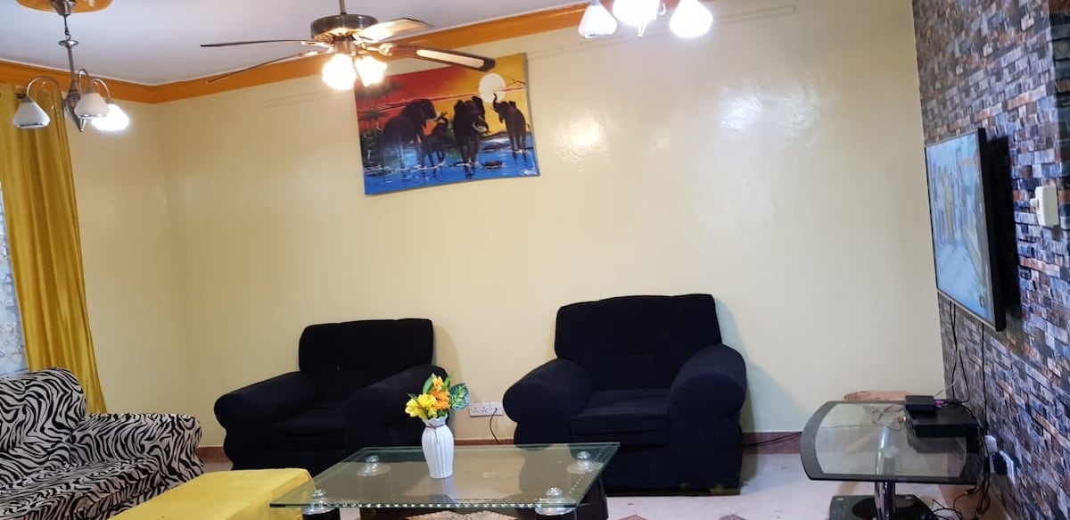 A sitting area features two dark sofas placed opposite each other, with a glass coffee table at the center. A flat-screen television is mounted on the wall, and decorative artwork depicting horses is displayed above. Soft yellow curtains frame a window, complementing the room's colors.