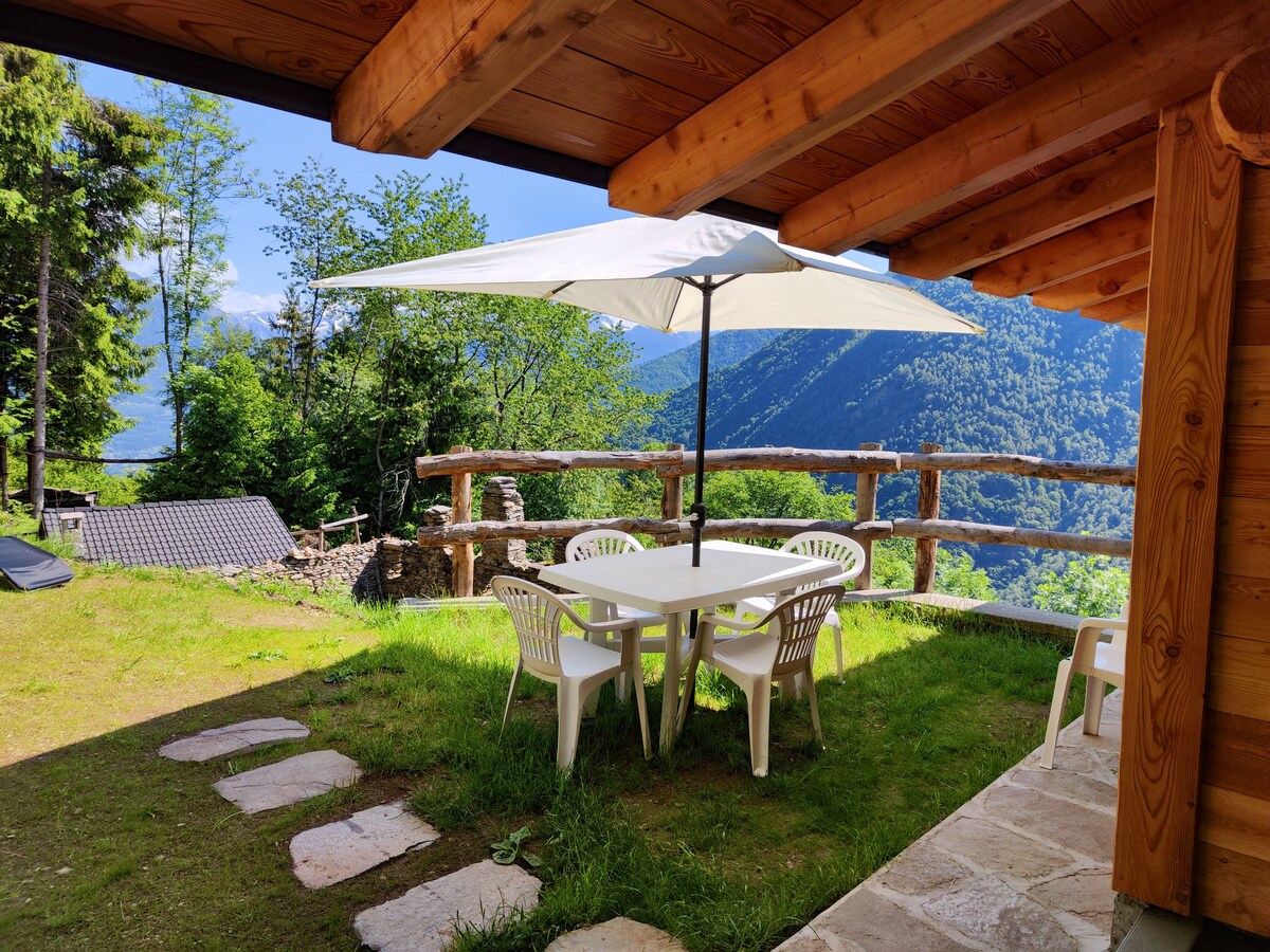 An outdoor dining area features a white table with four chairs, situated under a large parasol. A scenic view of lush greenery and distant mountains is visible in the background, enhancing the space's natural feel.