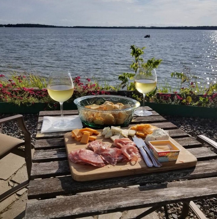 A wooden table is set by the lake, featuring an assortment of cheeses, sliced meats, and a bowl of fresh fruit. Two glasses of white wine stand beside the spread, with the serene lake and blooming flowers providing a scenic backdrop.
