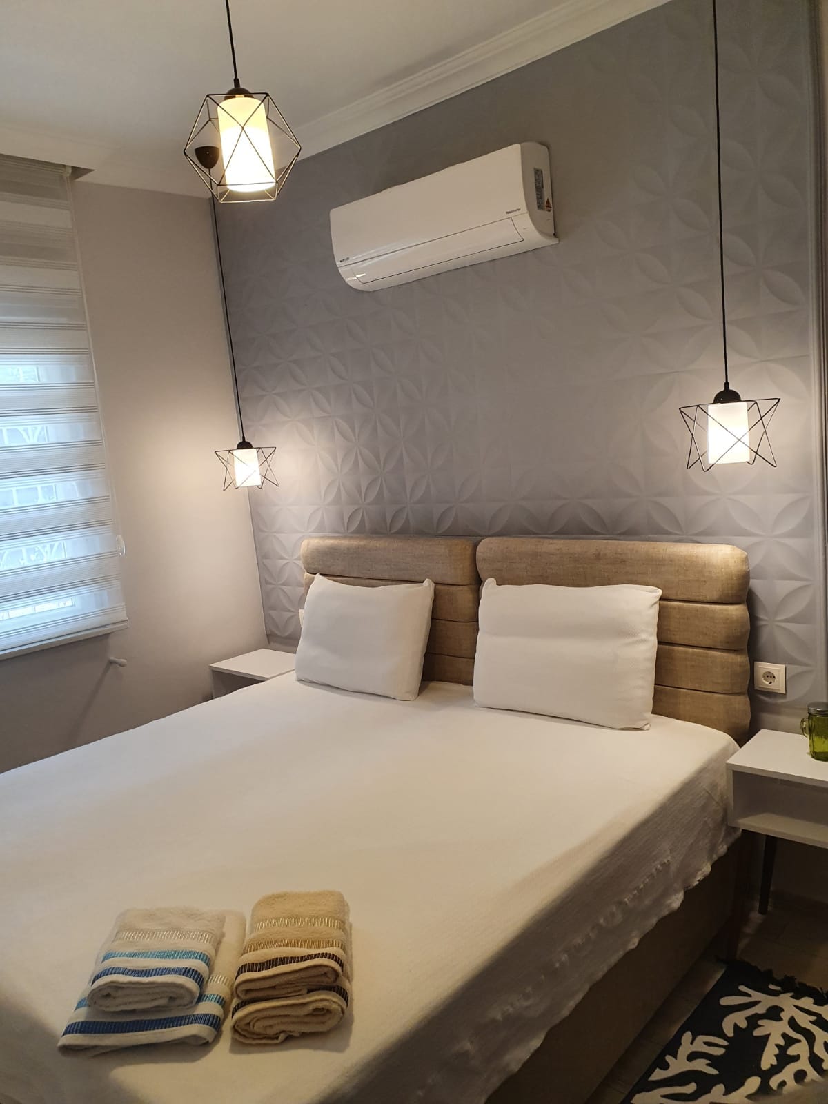 A cozy sleeping space features a spacious bed with light bedding and two neatly arranged towels at its foot. Soft lighting is provided by pendant fixtures above the bed, and a window draped with sheer fabric allows for natural light.
