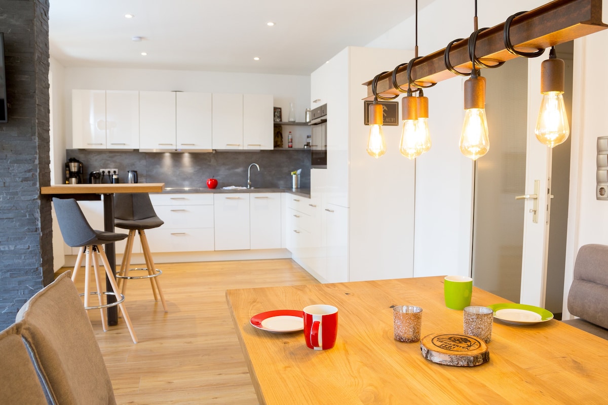 An open kitchen and dining area showcase minimalistic white cabinetry and a dark stone accent wall. A wooden dining table is set with colorful mugs, while pendant lights illuminate the space. Bar stools are positioned at the kitchen counter, inviting casual dining and interactions.
