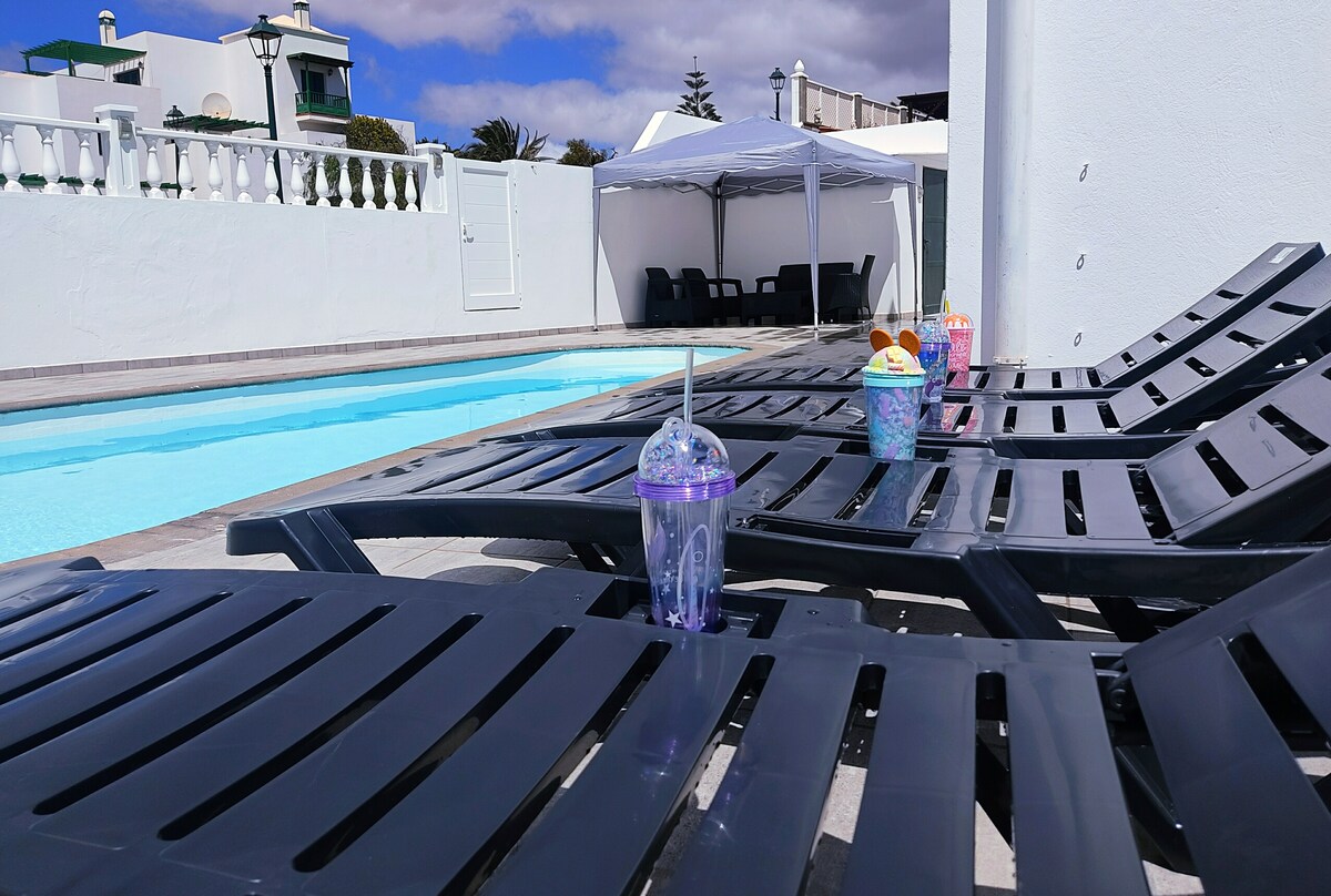 A row of black lounge chairs is situated beside a sparkling heated pool. Colorful drink cups are placed on the chairs, adding a vibrant touch to the serene outdoor space, which is bordered by white walls and shaded by a large tent.