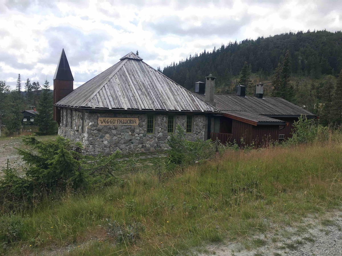 A traditional stone building with a distinctive round shape and a sloped roof is nestled in a natural setting. Surrounding greenery and trees complement the structure, which features a tall, pointed tower. The area is marked by grass and gravel, with a backdrop of gentle hills.