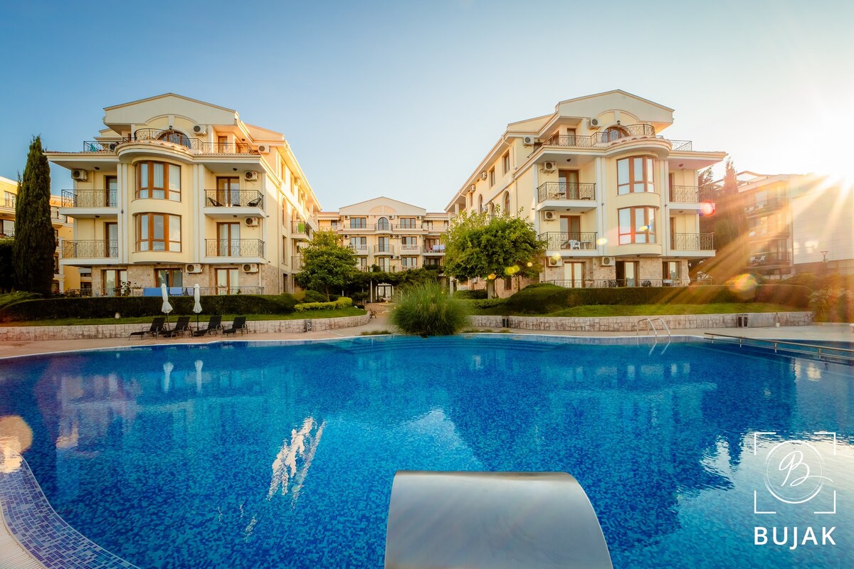 The image features a tranquil pool surrounded by well-maintained greenery and elegant buildings. The sun casts a warm light on the surface of the water, reflecting the architecture and enhancing the peaceful atmosphere of the complex.