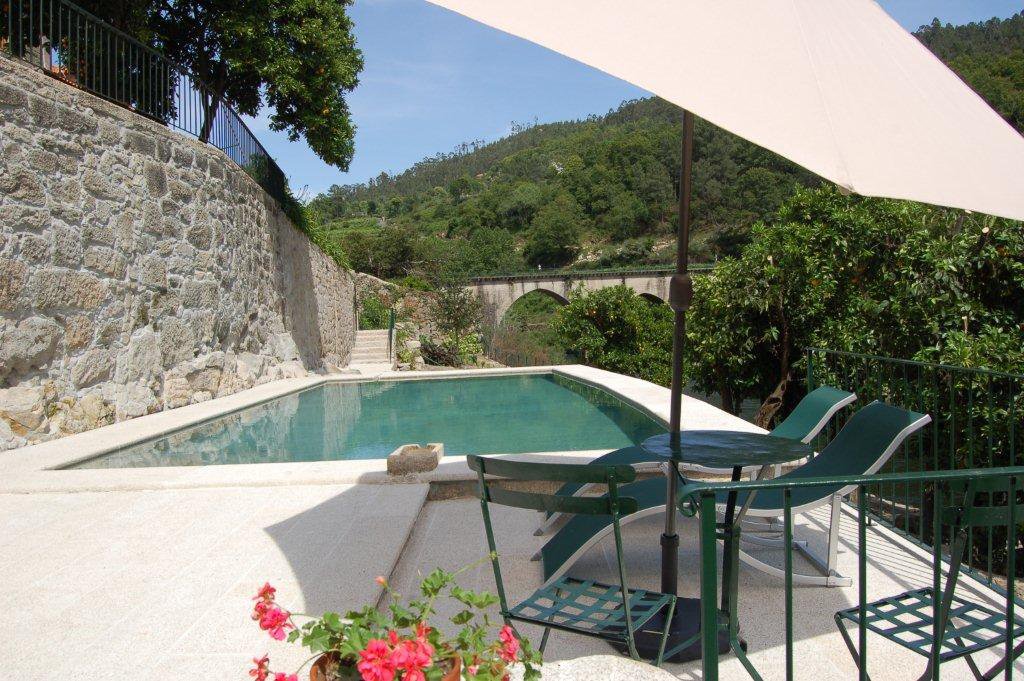 A clear swimming pool is set on a stone terrace surrounded by lush greenery. Lounge chairs are placed beside the pool, with a large umbrella providing shade. In the background, a picturesque stone bridge crosses the river, framed by rolling hills.