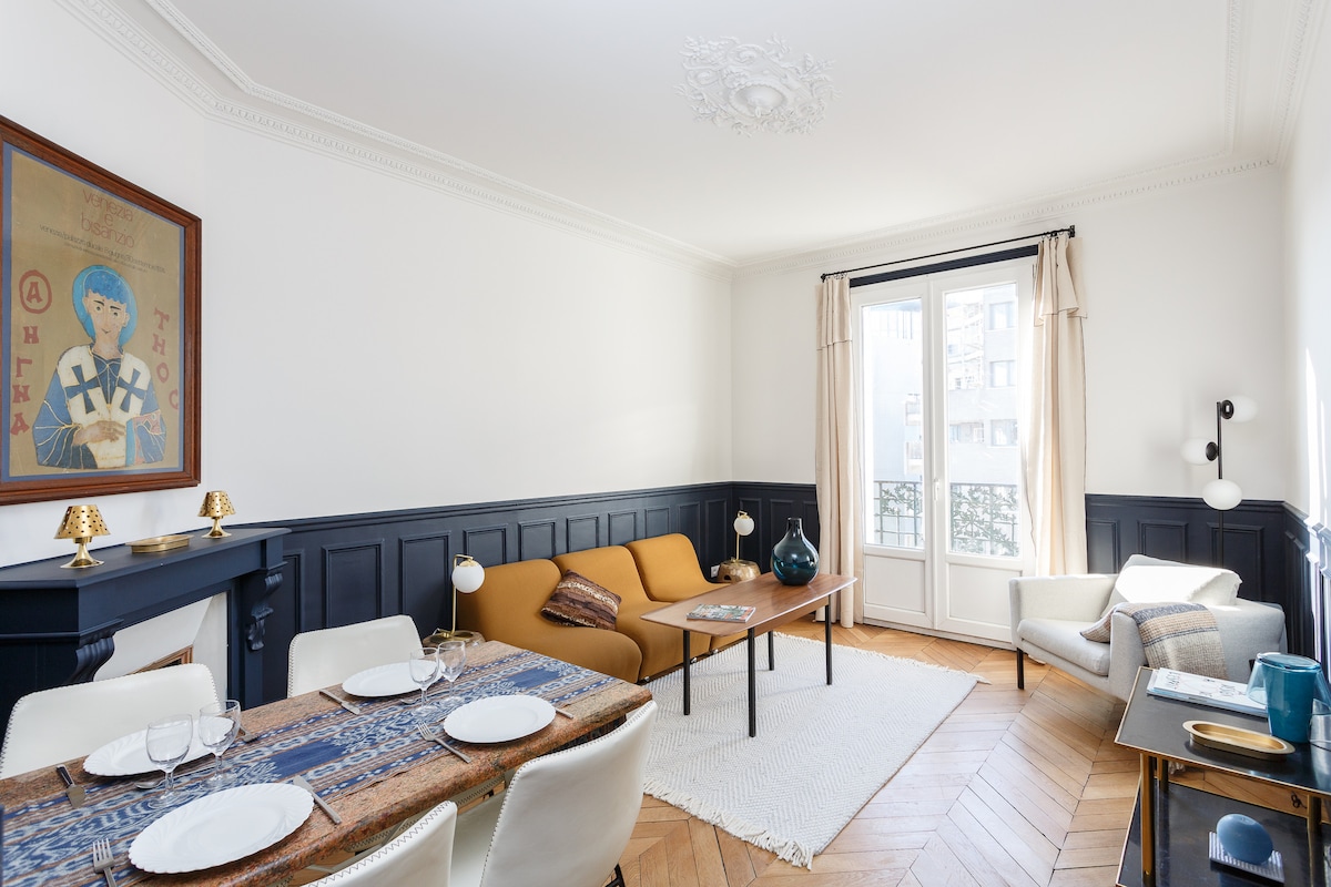 The living area features a combination of modern and traditional décor, including a sofa and a comfortable armchair. A dining table set for six is positioned in the foreground. Natural light enters through a large window, highlighting the contrasting colors of the walls and furnishings.