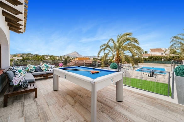 An outdoor terrace features a billiards table set against a backdrop of palm trees and mountains. Comfortable seating is arranged nearby, offering views of the private pool and landscaped yard. The clear sky adds to the serene atmosphere of the shared space.