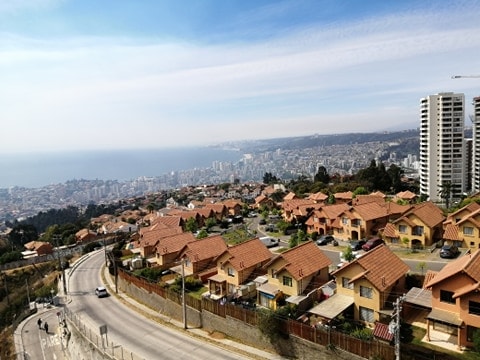 A panoramic view captures a sprawling hillside community with numerous homes featuring red-tiled roofs. Below, the coastline and cityscape of Viña del Mar can be seen, stretching towards the horizon under a clear blue sky with faint wisps of clouds.
