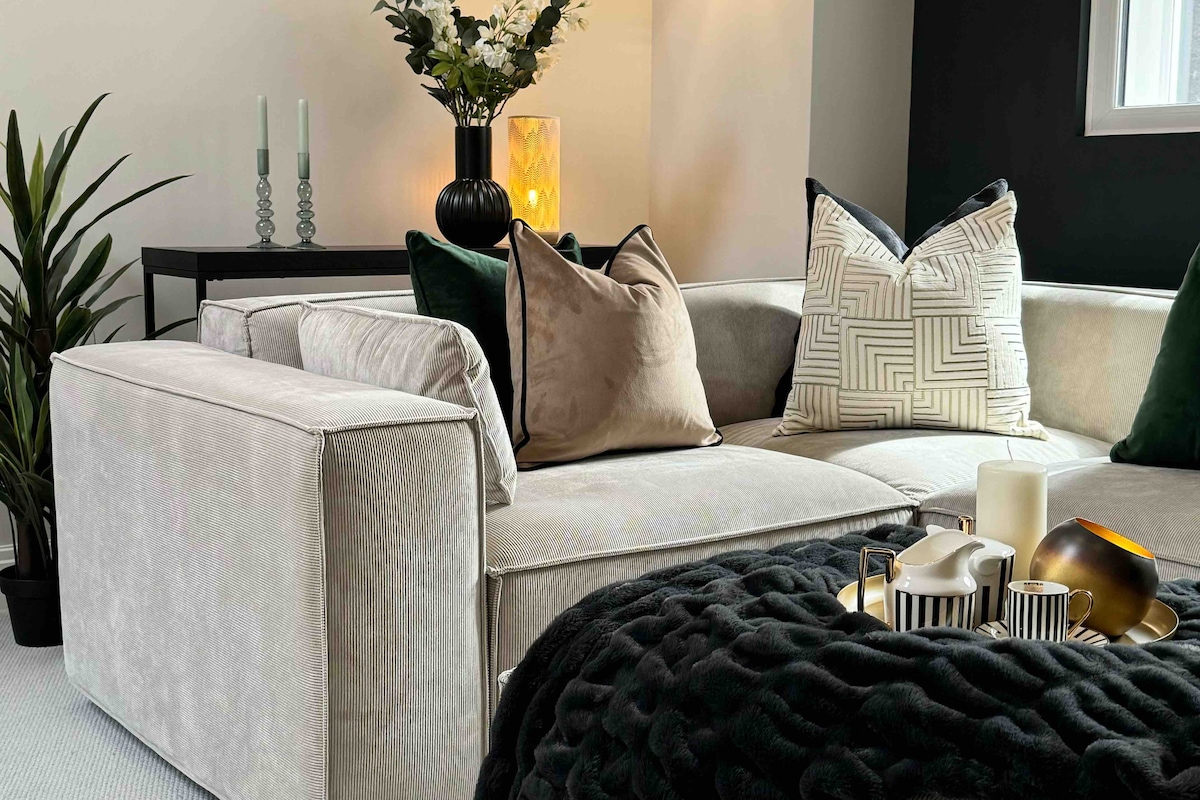 A spacious corner sofa is adorned with decorative pillows in various textures and patterns. A small coffee table holds an arrangement of candles and dishware. Subtle greenery is present nearby, alongside a console table with decorative accents.