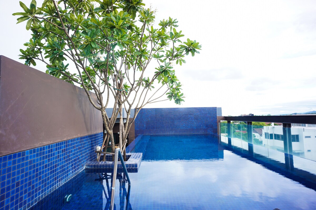 A roof-top pool is showcased, featuring deep blue tiles and a tranquil water surface. A single tree is positioned nearby, adding a touch of greenery. The surrounding area offers unobstructed views of the sky and cityscape.
