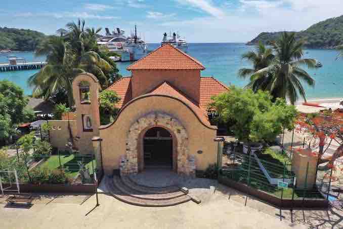A charming building with a terracotta-tiled roof is situated near a sandy beach, framed by palm trees. The ocean is visible in the background, with cruise ships docked at the pier. Lush greenery surrounds the entrance, adding to the serene coastal ambiance.
