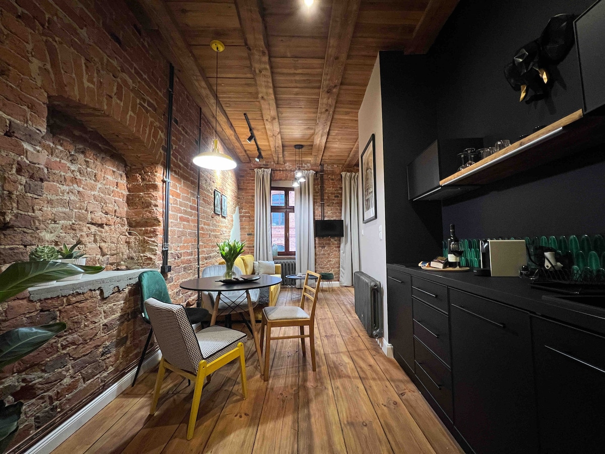 A modern dining area is seen featuring a round wooden table surrounded by six chairs. Exposed brick walls complement the wooden ceiling, while large windows allow natural light to brighten the space. A sleek kitchen with matte black cabinetry is visible in the background.