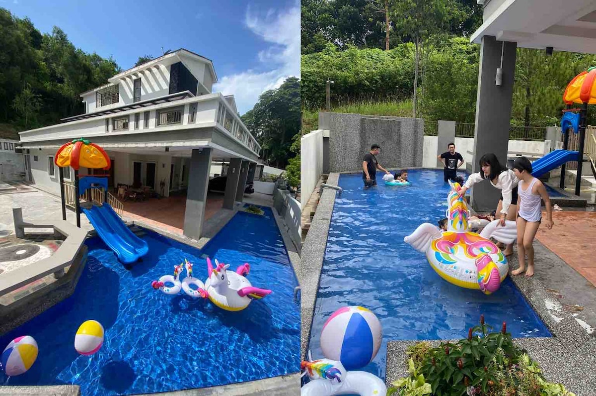An outdoor kids swimming pool is shown, featuring a slide and colorful inflatable toys. A spacious villa is visible in the background, with a covered patio area. The pool area is surrounded by a fence and lush greenery, providing a vibrant and playful environment for families.