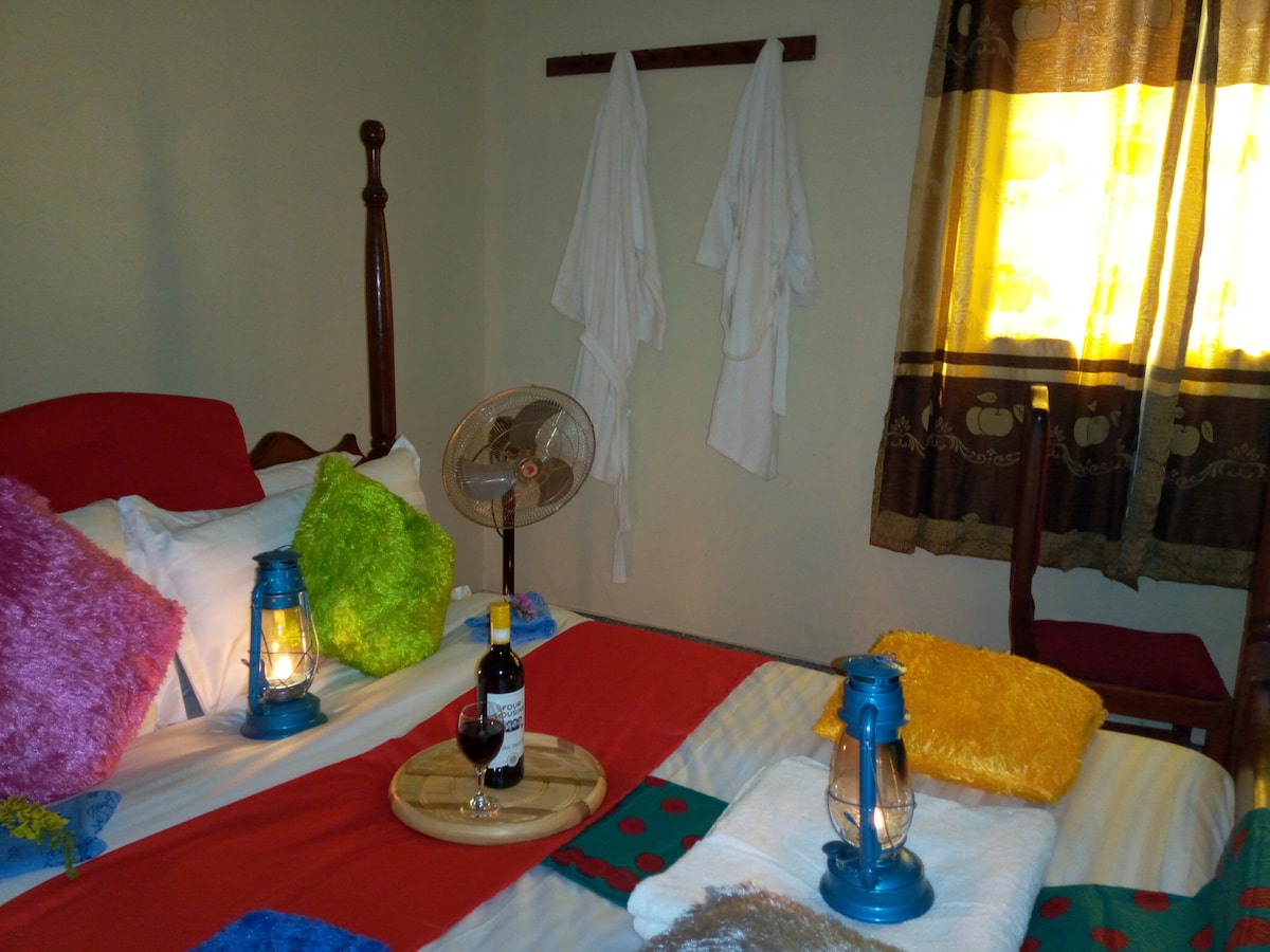 A comfortable sleeping area features a spacious bed adorned with colorful decorative pillows. Soft lighting is provided by two lanterns on the bedside, accompanied by a bottle of wine and a glass. A ceiling fan offers a cool breeze, while two robes hang nearby. Natural light filters through a partially drawn window.